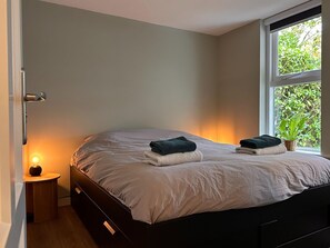 A bedroom with a bed, a bedside table with a lamp, a window with a view of greenery, and a potted plant on the windowsill.
