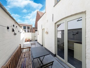 A narrow courtyard with a wooden bench, a table, and chairs. There is a white wall with a string of lights and a door leading to a garage. - Luxurious Holiday Home in Nieuwpoort-stad (Nieuwpoort)