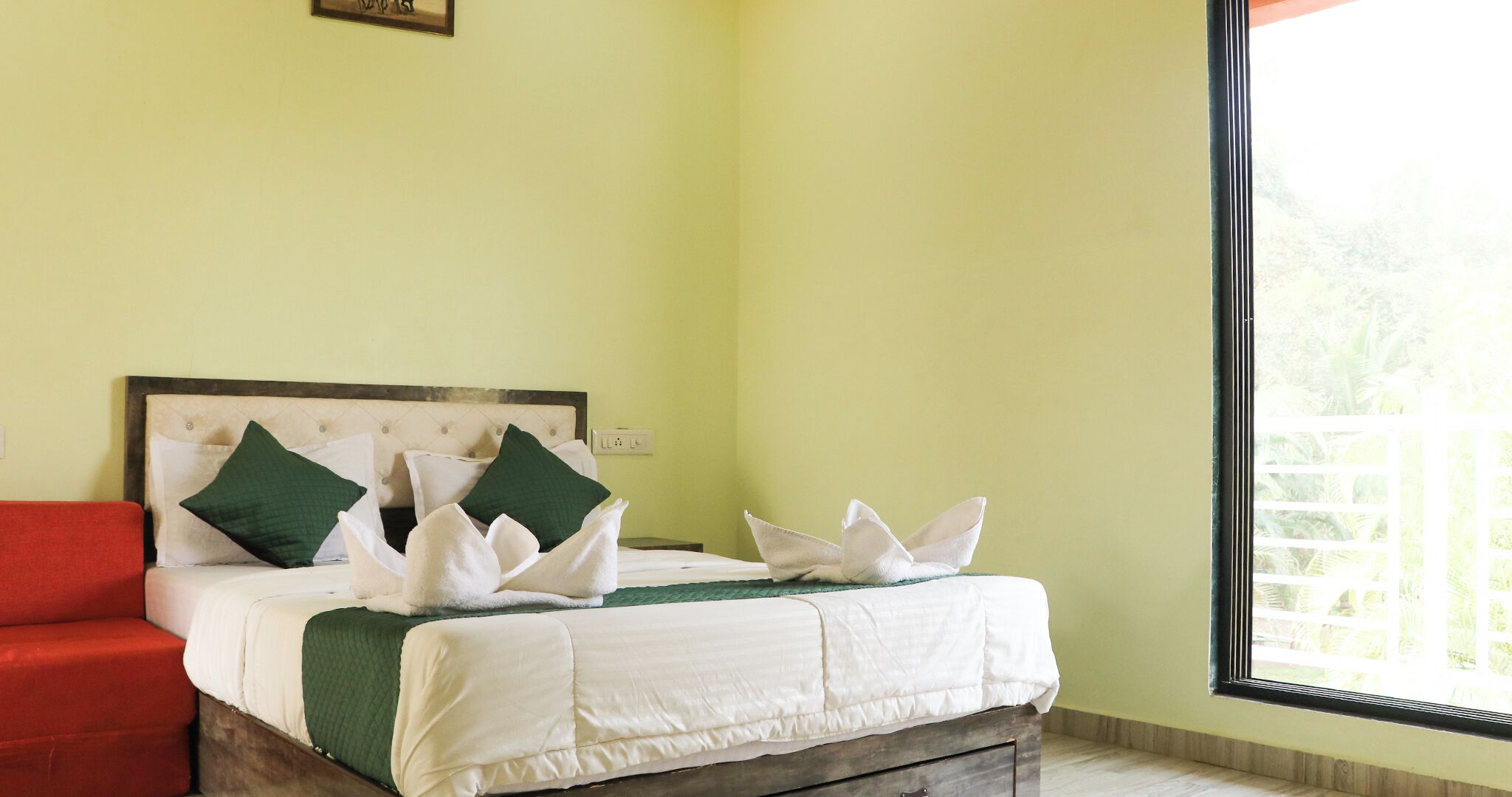 A bed with white linens and green pillows, a red chair, and a window with a view of greenery.