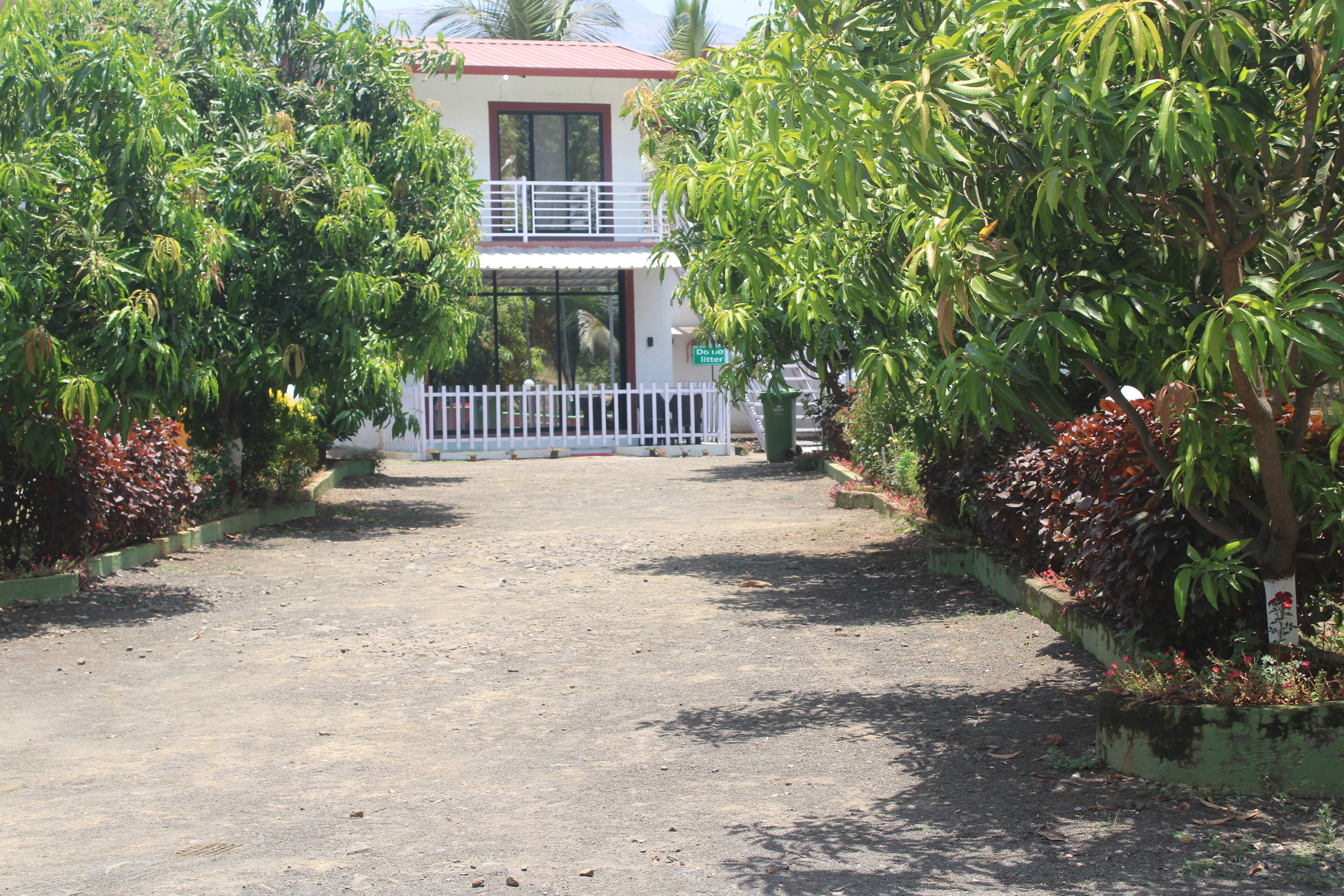A two-story building with a white exterior and a red roof. There is a white fence in front of the building, and the area is surrounded by lush greenery and plants.