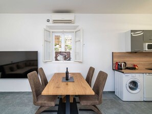 A compact living space with a wooden dining table, four chairs, a TV, a window with blinds, a microwave, and a washing machine. - Duxperience Apartment (Dubrovnik)