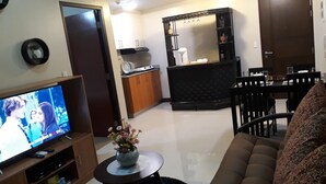 A living room with a television, a sofa, a dining table with chairs, a kitchen area, and a large mirror.