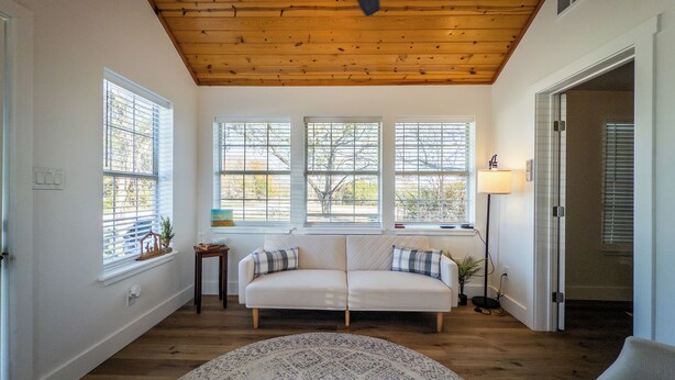 A spacious room with a wooden ceiling, a white sofa with patterned cushions, a round rug, and large windows with blinds.