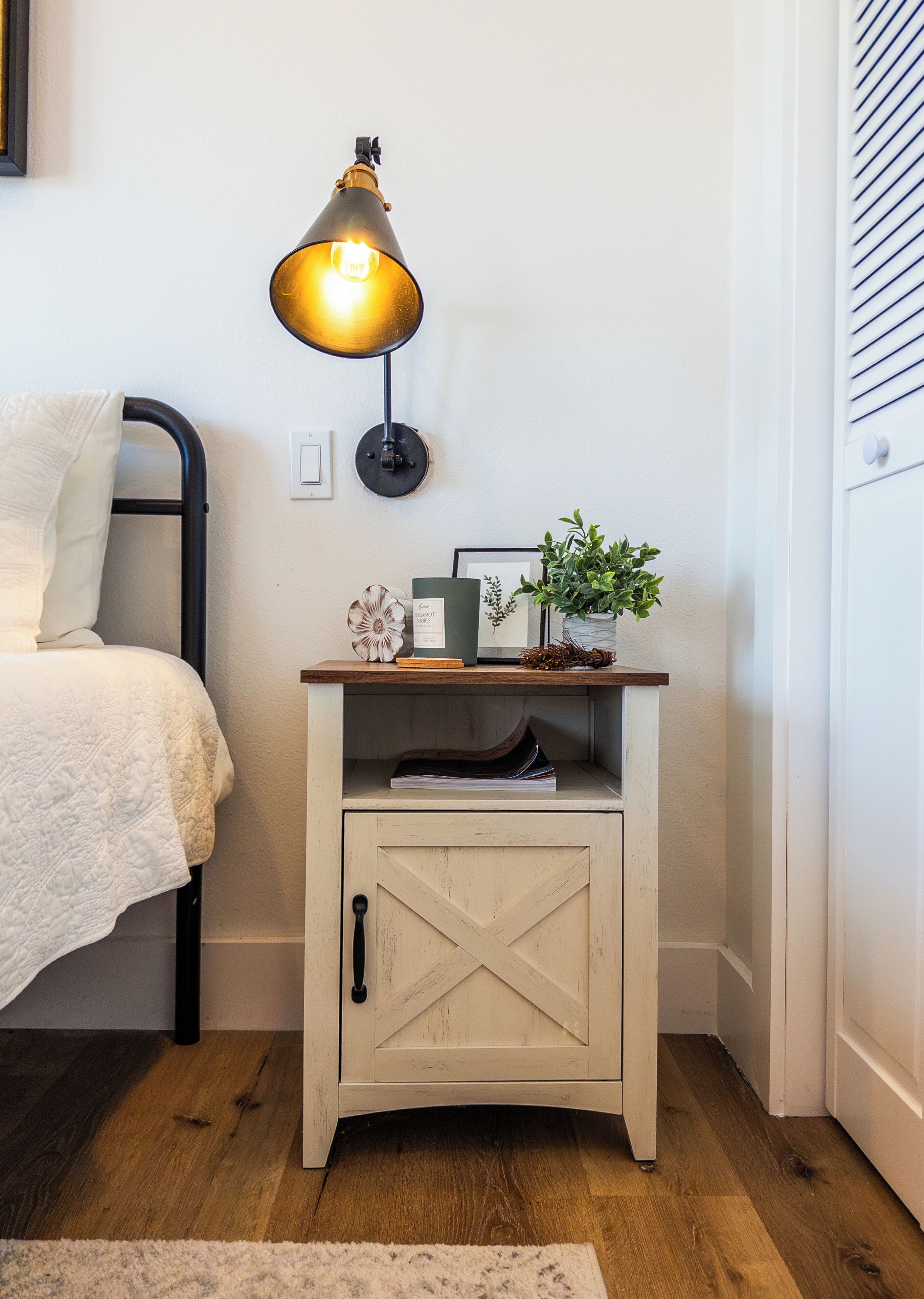 Een slaapkamer met een bed, nachtkastje met een lamp, een klein kastje en een wandlamp.