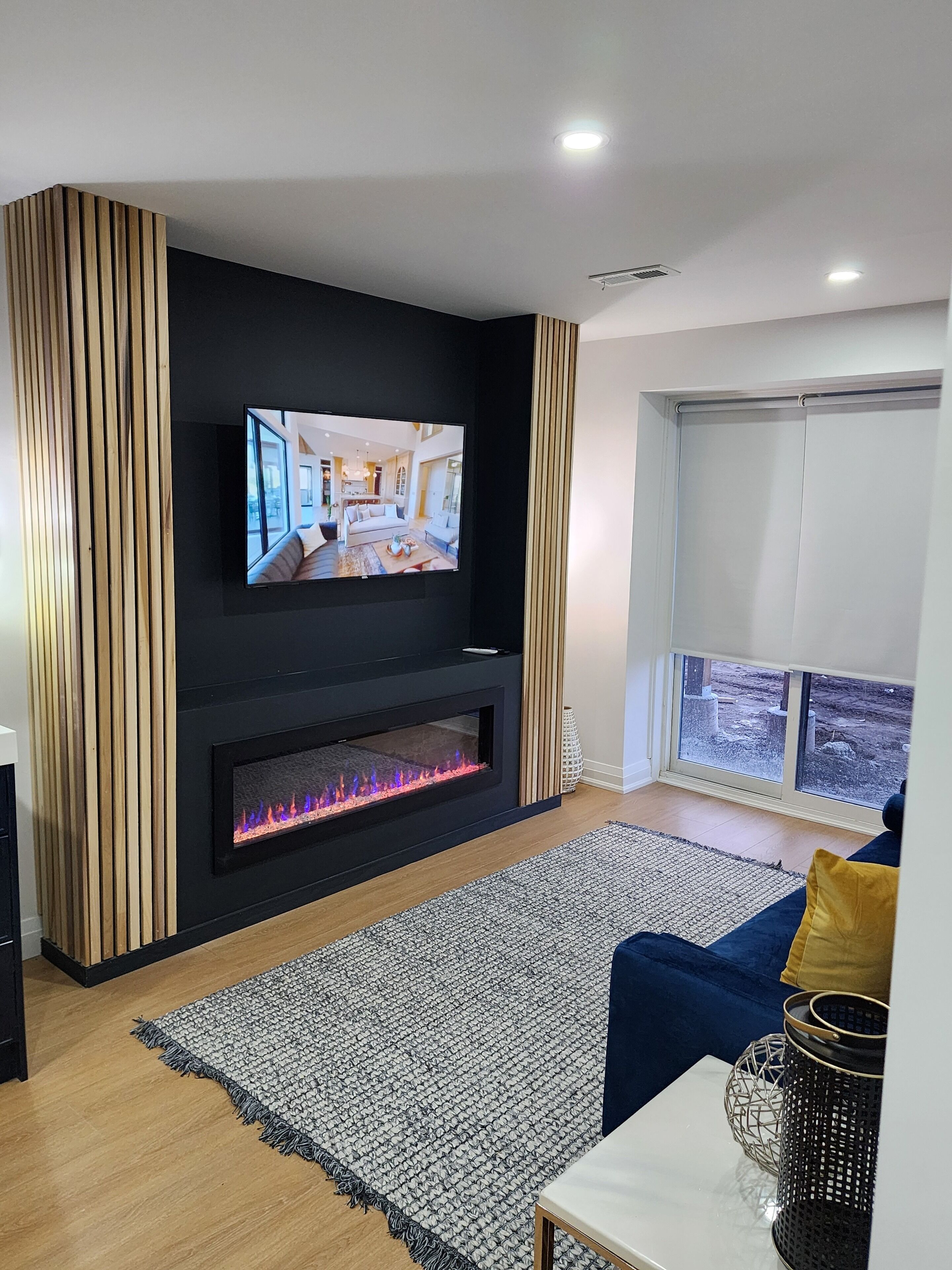 A modern living room with a flat-screen TV mounted on a black wall, a fireplace with a glass front, and a large window offering a view of the outdoors.