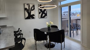 Een eetgedeelte met een ronde tafel, zwarte stoelen en een potplant. Er hangen abstracte schilderijen aan de muur en er is uitzicht op een bouwplaats door het raam.