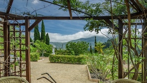 A covered outdoor seating area with wicker chairs and a mosaic-tiled table, overlooking a landscaped garden with various plants and a distant mountain view.