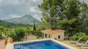 A swimming pool with a tiled deck, surrounded by greenery and a mountain in the background.