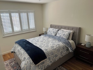 Une chambre à coucher comprenant un lit, deux tables de chevet, une fenêtre avec des stores et une petite table de nuit avec une lampe.