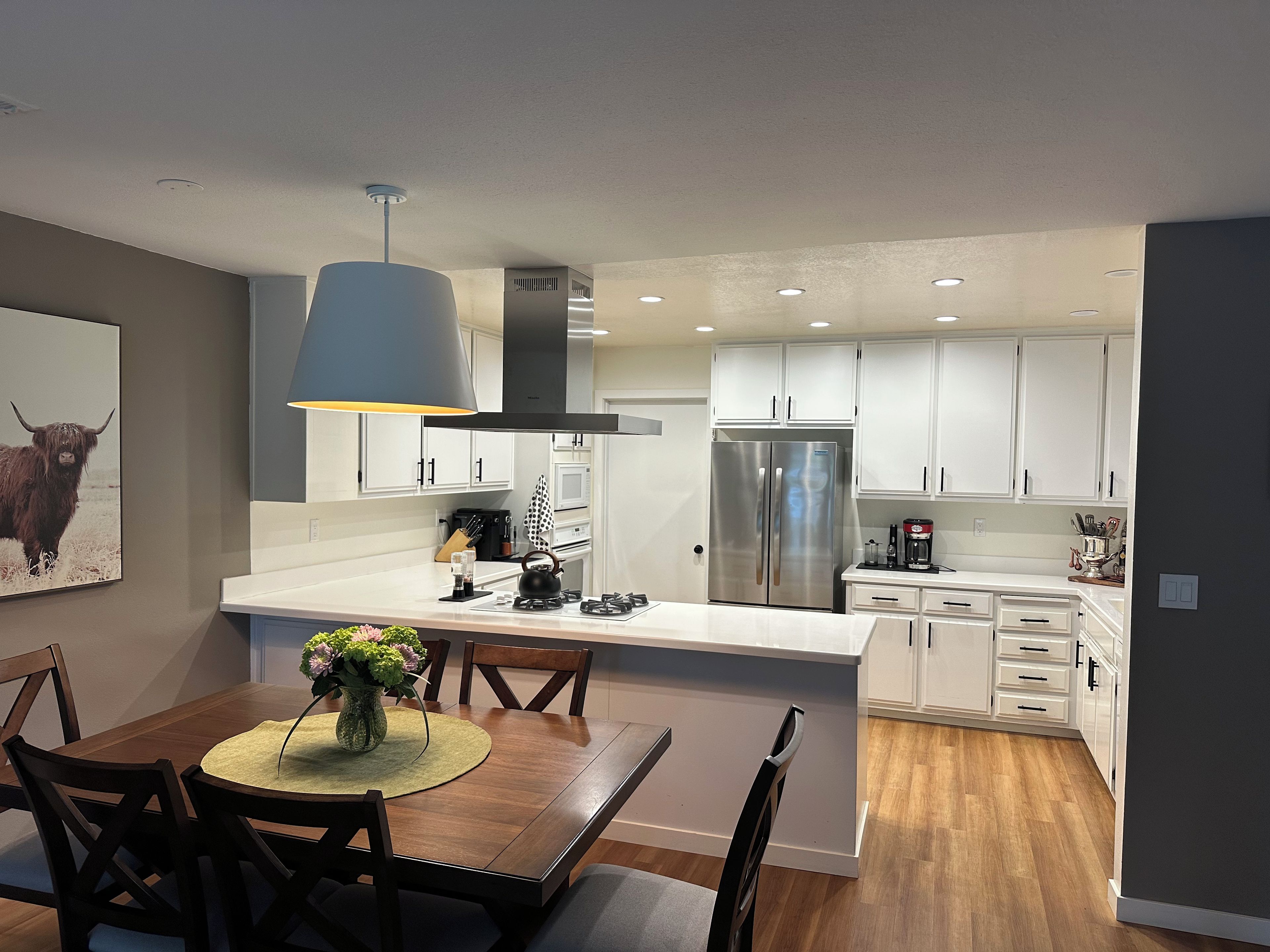 A modern kitchen with white cabinets, a stainless steel refrigerator, and a dining area with a wooden table and chairs.