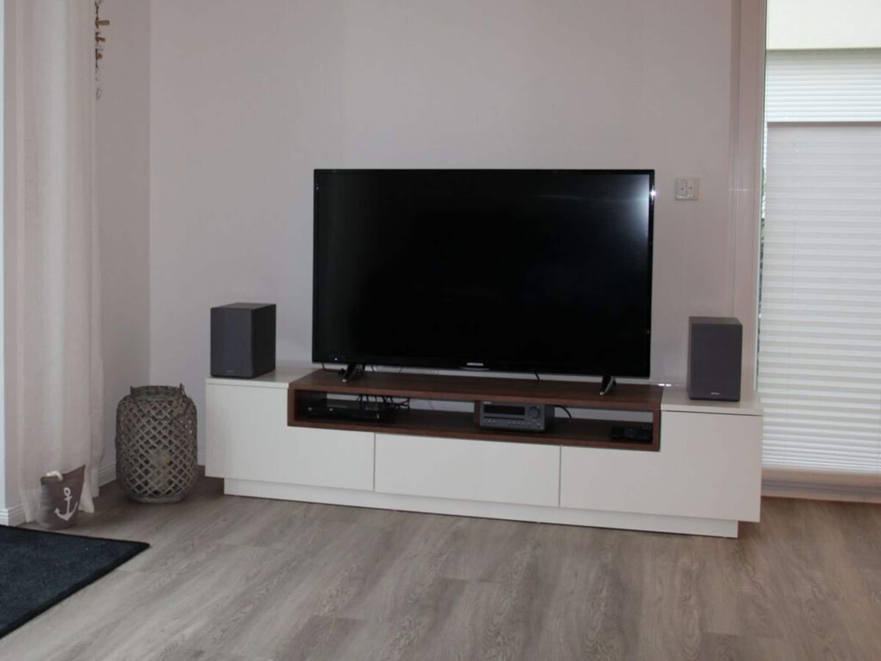 A modern living room with a flat-screen TV on a wooden stand, two speakers, and a small shelf with electronic devices.