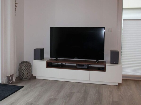 A modern living room with a flat-screen TV on a wooden stand, two speakers, and a small shelf with electronic devices. - Holiday Home Kellenbude (Kellenhusen)