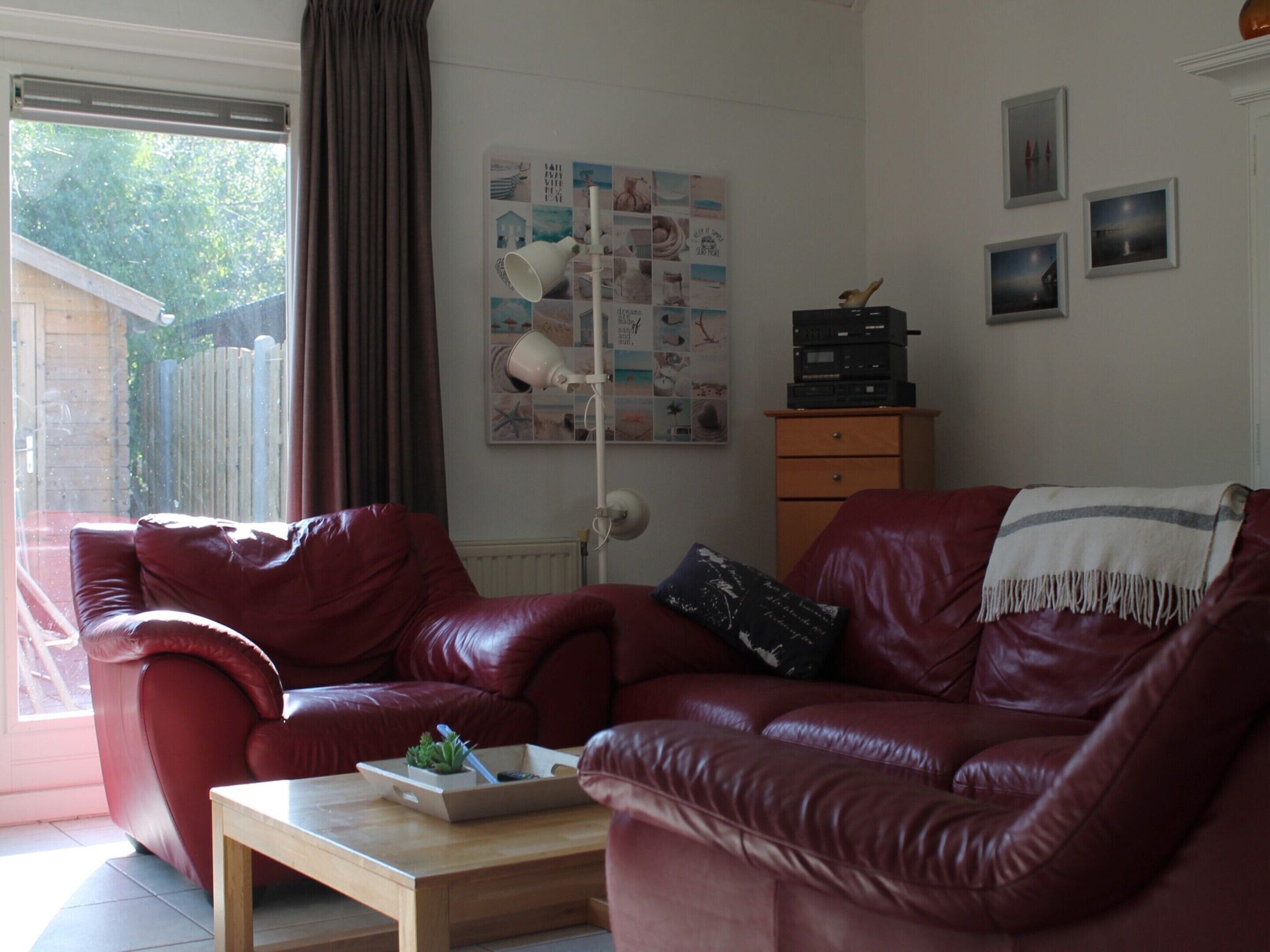 A living room with a red leather sofa, a wooden coffee table, and a radiator. There is a window with curtains, a wall with framed pictures, and a small cabinet with a radio.