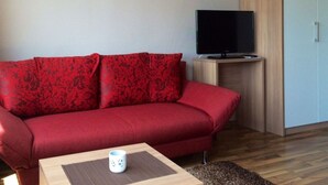 A red sofa with floral upholstery, a television on a wooden stand, and a small white mug on a wooden table. - Saaleland Apartment 3 (Schleiz)