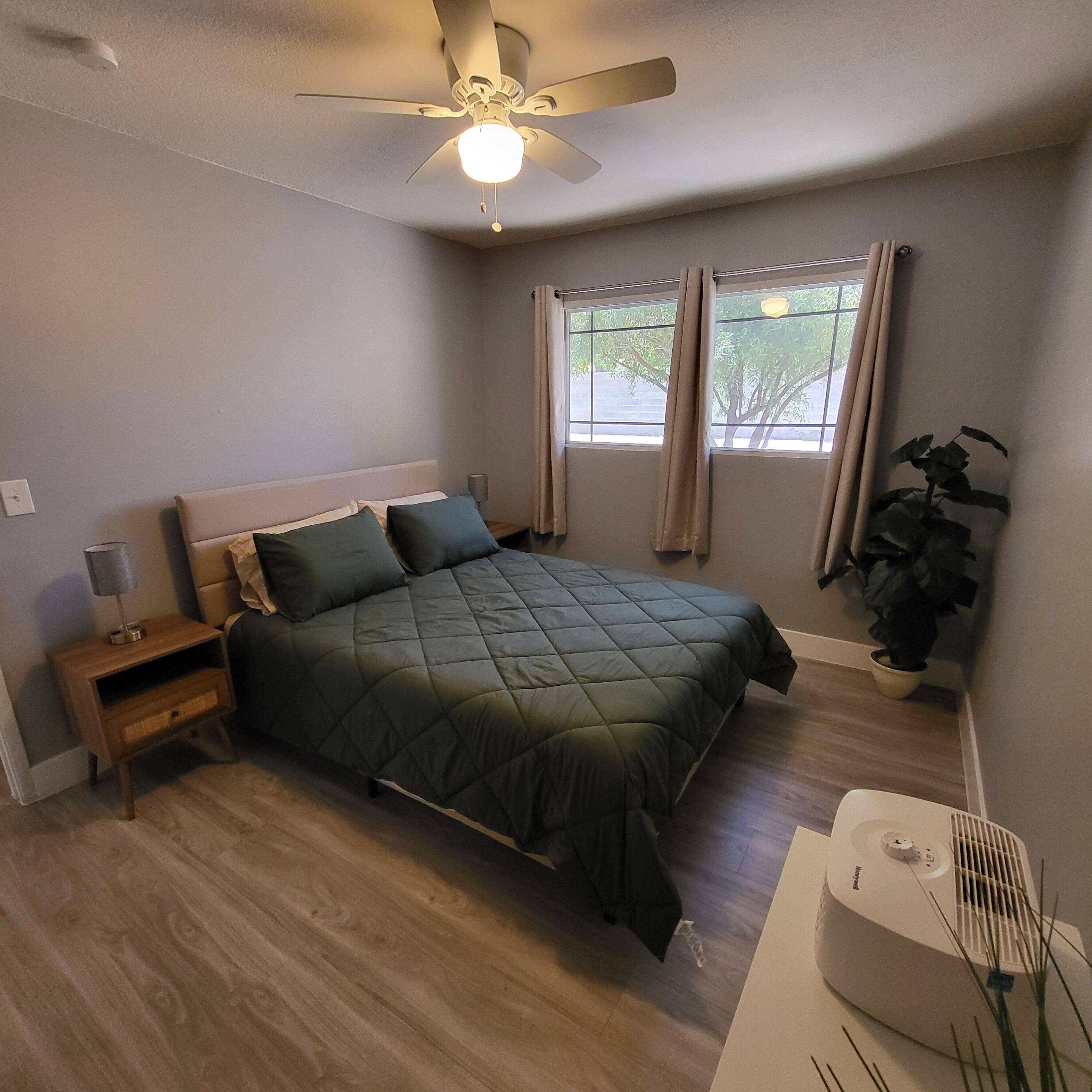Quarto com cama, mesa de cabeceira, ventoinha no teto, janela com cortinas e um vaso com planta.