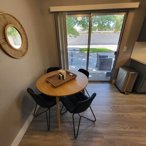 Un comedor con una mesa redonda, cuatro sillas, un espejo y vistas a un espacio exterior con una calle y vegetación a través de un ventanal.