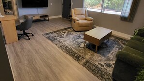 Ein Wohnzimmer mit Flachbildfernseher, Deckenventilator, beigem Sessel, Couchtisch und großem Fenster mit Blick auf Grünflächen.