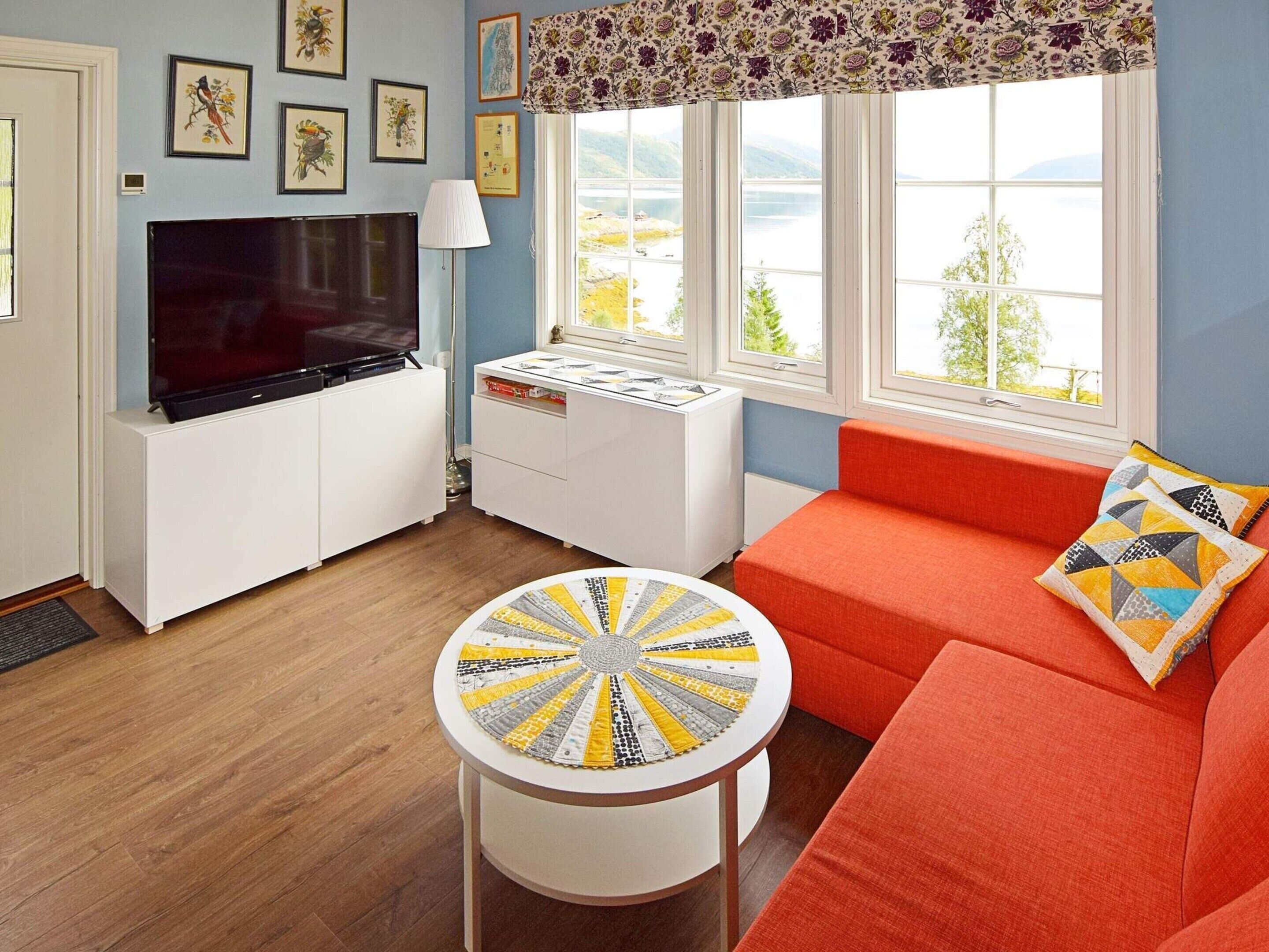 A living room with a TV, a round table, a sofa, and a window with a view of a lake.