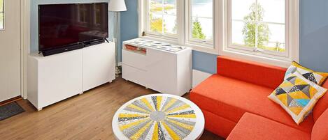 A living room with a TV, a round table, a sofa, and a window with a view of a lake.