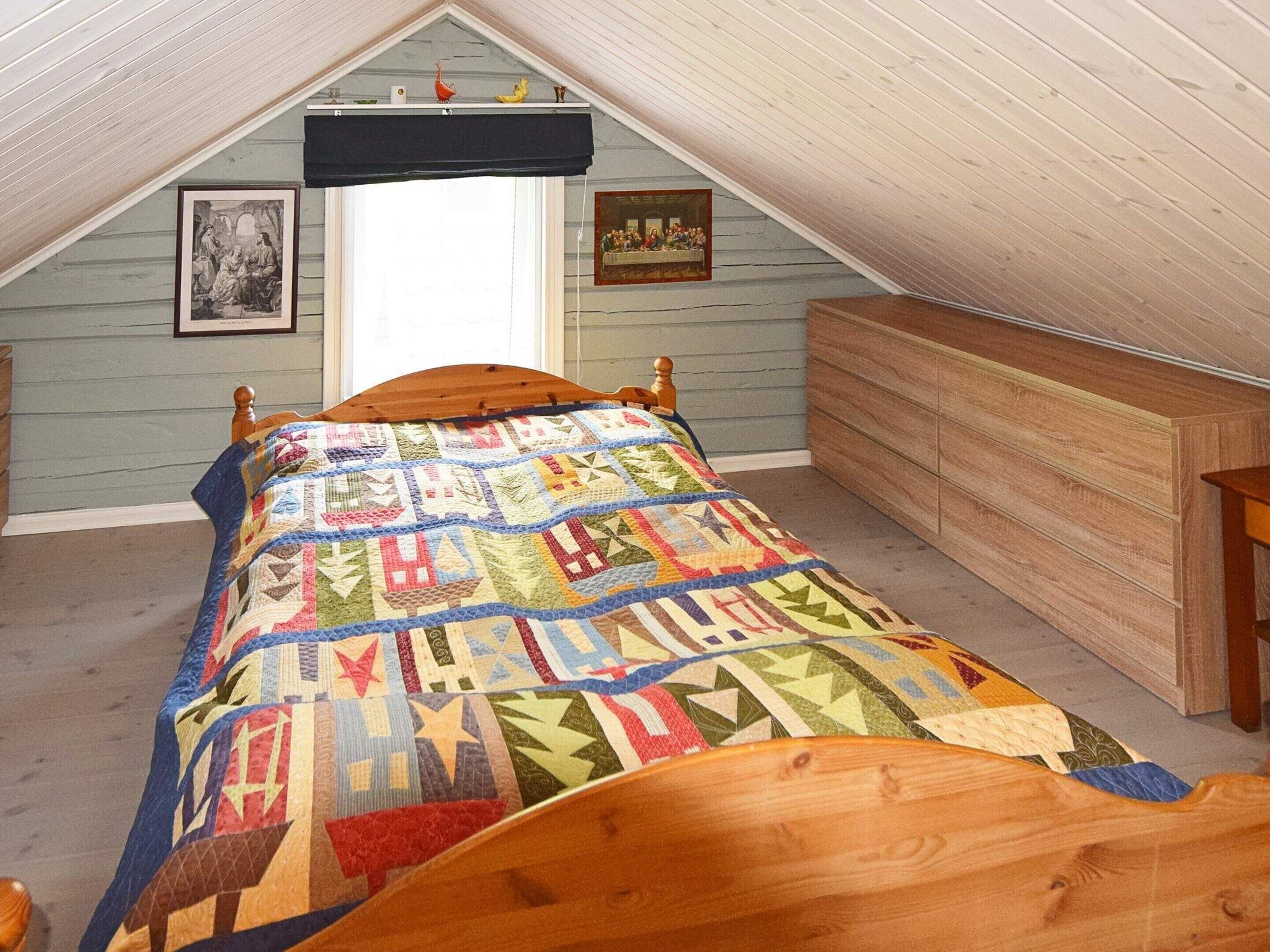 A bedroom with a sloped ceiling, a wooden bed with a colorful quilt, a wooden dresser, and two framed pictures on the wall.