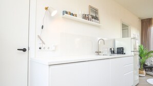Une cuisine moderne avec des armoires blanches, un évier encastré et une machine à café. Une étagère se trouve au-dessus de l’évier, où sont disposés des objets décoratifs et un tableau encadré.