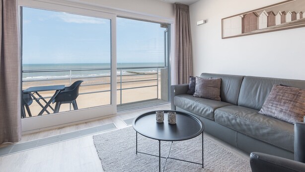 Un salon moderne avec un canapé gris, une petite table ronde et une vue sur la plage grâce à de grandes portes-fenêtres coulissantes.