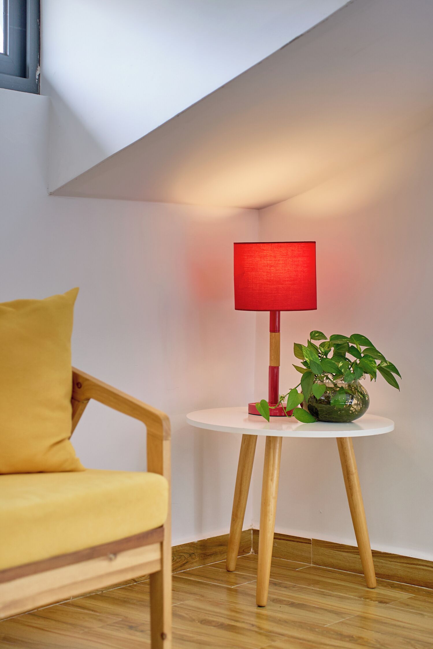 Een hoek van een kamer met een houten stoel, een witte bijzettafeltje met een rode lamp en een potplant.