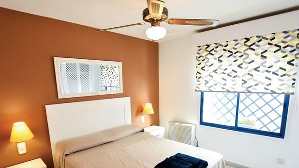 A bedroom with a bed, bedside lamps, a ceiling fan, a window with patterned blinds, and a radiator. - Belvilla by OYO Minarete 10A (Roquetas de Mar)