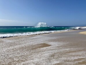 Une plage où les vagues déferlent sur le rivage, un ciel dégagé et quelques personnes se promenant le long de la plage.