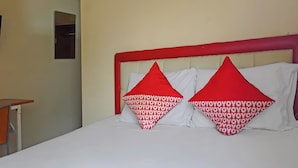 Een hotelkamer met een bed, twee rode en witte kussens, een wit hoofdeinde en een klein tafeltje met een stoel.