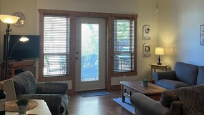 A living room with a sofa, a coffee table, a TV, and a lamp. There are two doors leading outside. - Lakefront Home away from Home (Osoyoos)