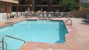 A swimming pool with a white fence, lounge chairs, and a building with balconies in the background. - Lakefront Home away from Home (Osoyoos)