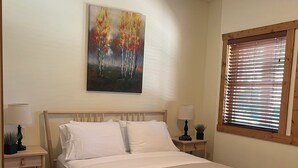 A neatly arranged bedroom with a bed, bedside tables, a ceiling fan, a window with blinds, and a painting on the wall. - Lakefront Home away from Home (Osoyoos)