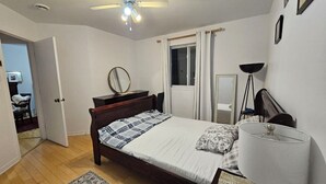A bedroom with a bed, a bedside lamp, a window with curtains, a ceiling fan, and a door leading to another room.