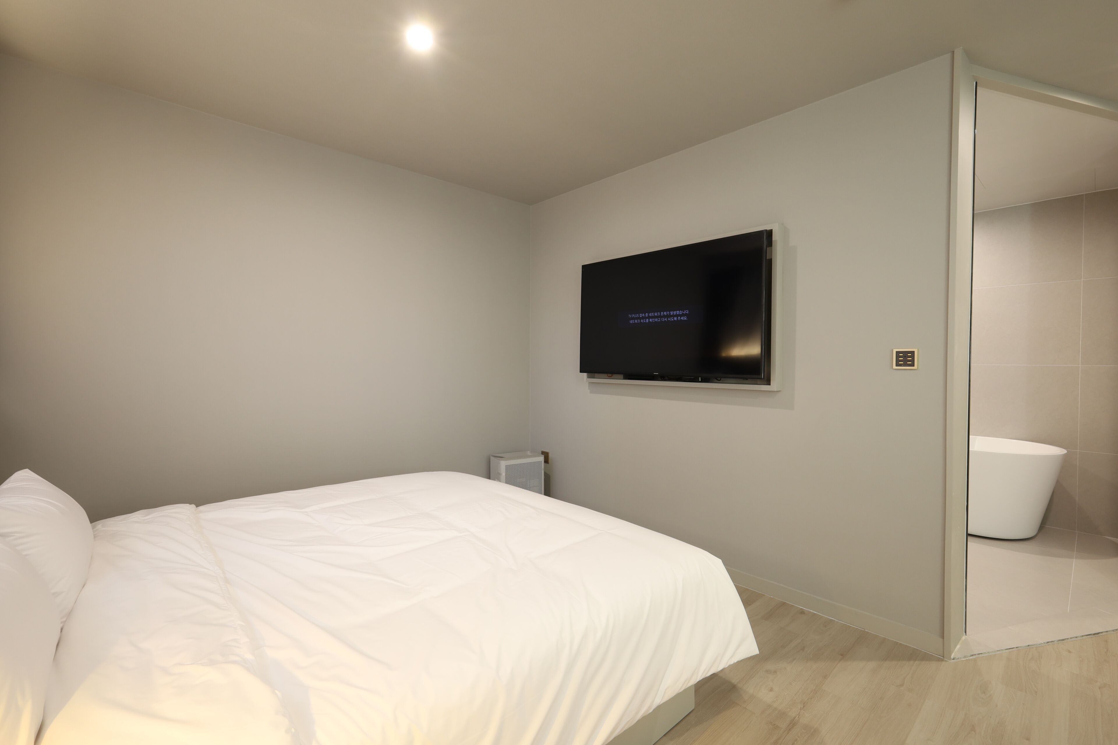 A modern hotel room with a large bed, a flat-screen TV mounted on the wall, and a glass partition separating the room from the bathroom.