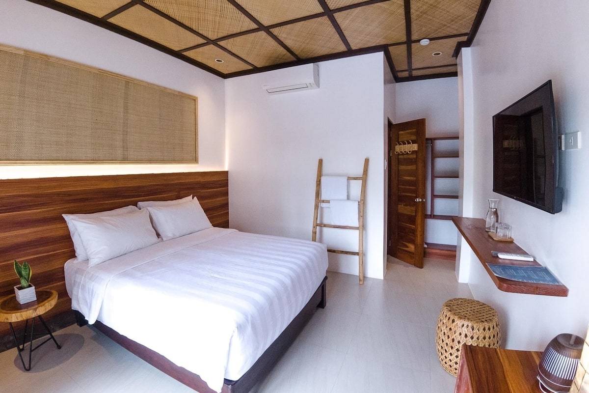 Un dormitorio moderno con una cama grande, cabecera de madera, televisor montado en la pared, una mesita de noche con una planta y un taburete tejido.