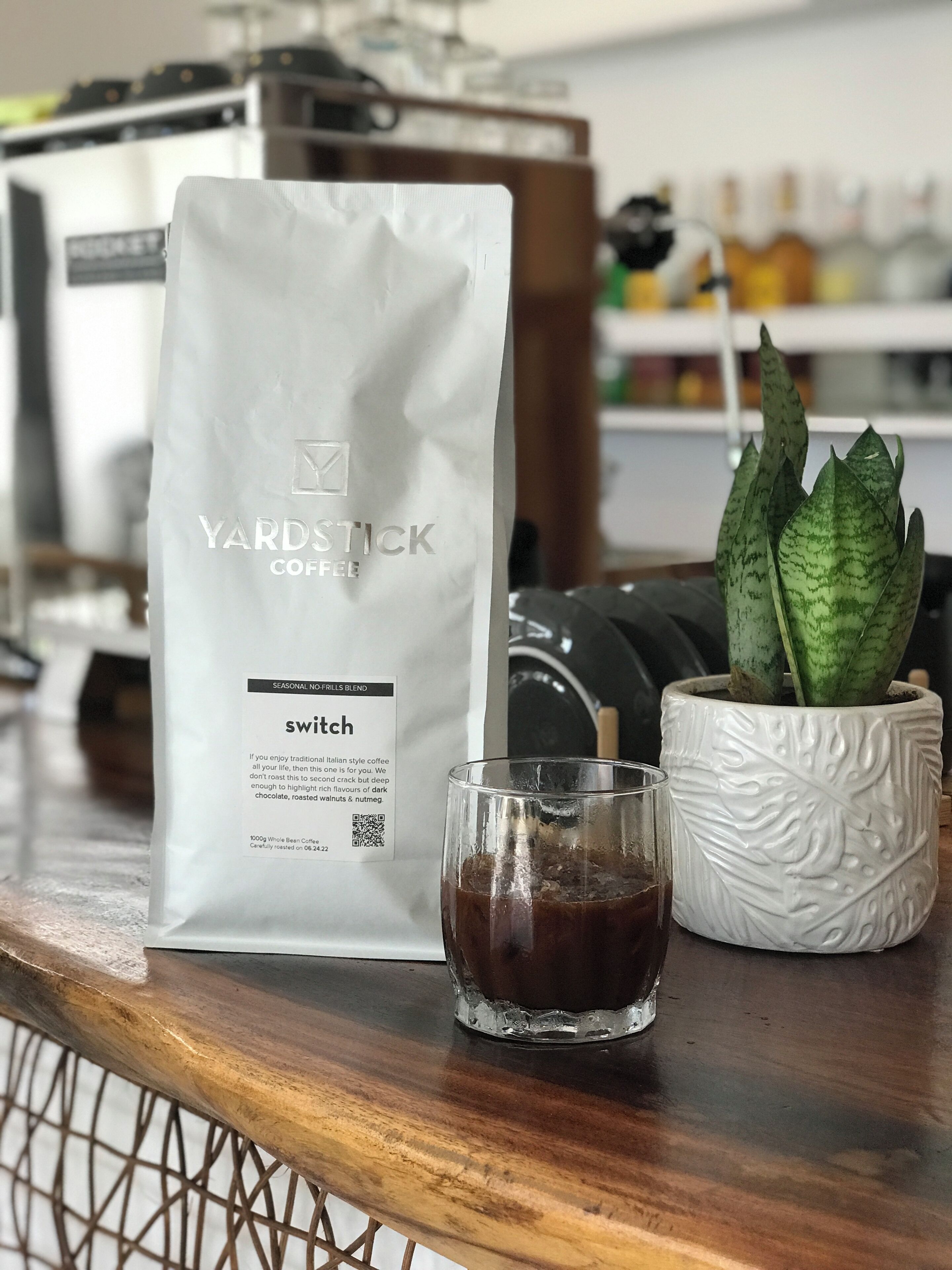 A coffee bag labeled 'Yardstick Coffee' next to a glass of coffee and a potted plant on a wooden surface.