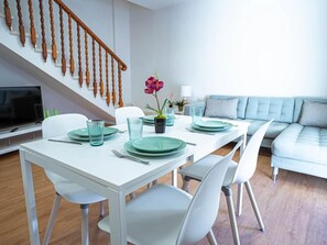 A dining area with a white table set for four, surrounded by white chairs, a staircase with wooden balusters, and a light blue sofa in the background. - Belvilla by OYO Apartment in Arona (Arona)