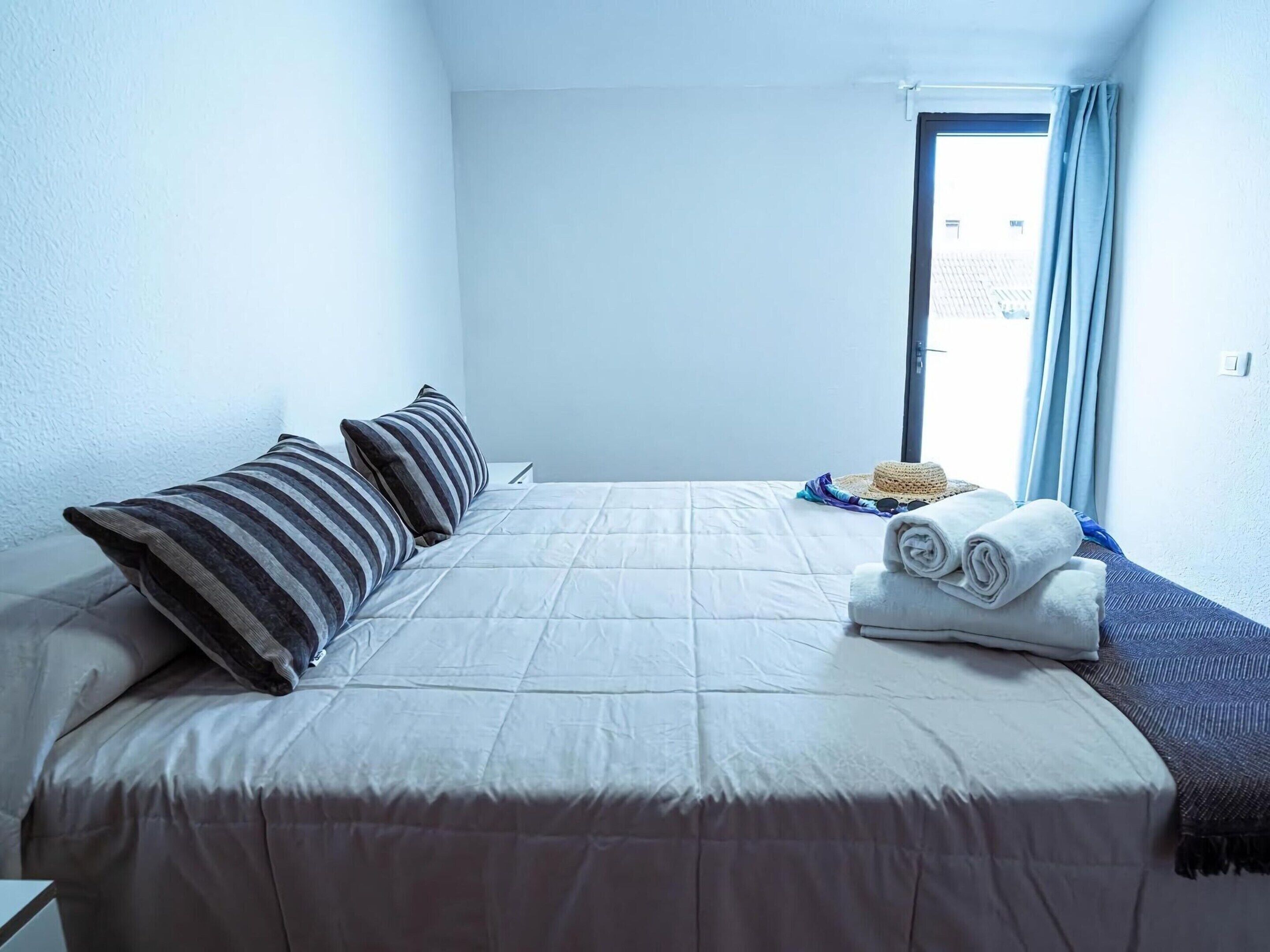 A hotel room with a bed, pillows, a rolled towel, a straw hat, and a window with curtains.