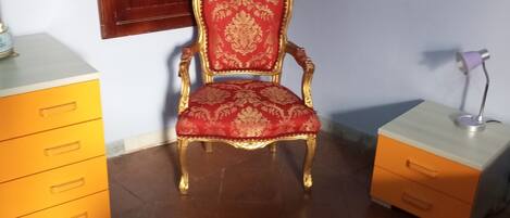 A room with a red upholstered chair with gold trim, a wooden cabinet with drawers, and a small bedside table with a lamp.