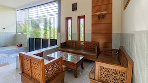 A spacious living area with a sofa set, a coffee table, and a TV. There are two windows with decorative glass panes and a wooden slatted wall. - SPOT ON 93965 Ibrahim Guest House Syariah (Ngasem)