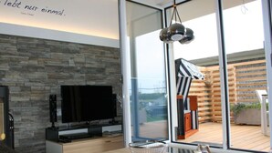 Sala de estar moderna com parede de pedra, TV de tela plana, mesa de centro de madeira e uma grande janela com vista para um deck de madeira e móveis de exterior.