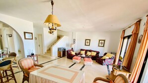 Amplia sala de estar con una mesa central, sillas de mimbre, un conjunto de sofás y una escalera que conduce a otro nivel.