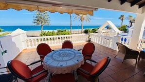 Una terraza con una mesa redonda y sillas rojas, con vistas a una playa con palmeras y el mar.