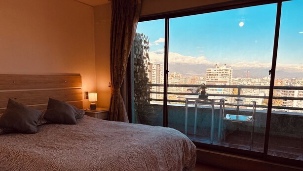 Een slaapkamer met een groot bed, een nachtkastje, een lamp en uitzicht op de stad vanaf een balkon.