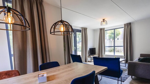 A modern hotel room with a wooden dining table, blue chairs, a grey sofa, and large windows with curtains. - Detached Holiday Home in Nijmegen With Sauna (Ewijk)