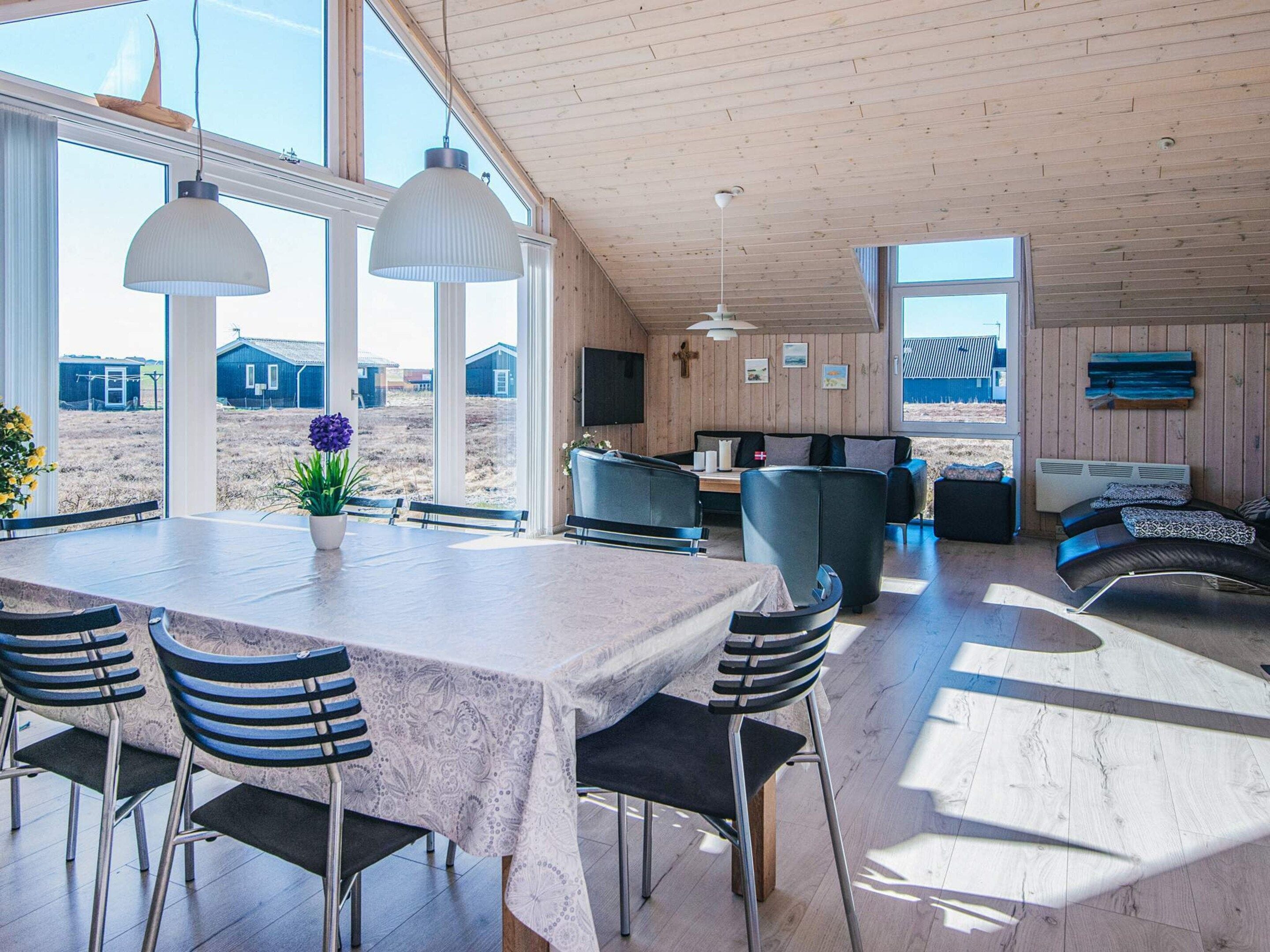 A spacious dining area with a large table and chairs, a kitchen area with a sofa, and large windows offering views of the surrounding landscape.