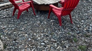 A table with four red Adirondack chairs on a rocky beach with a view of a calm body of water and distant land.