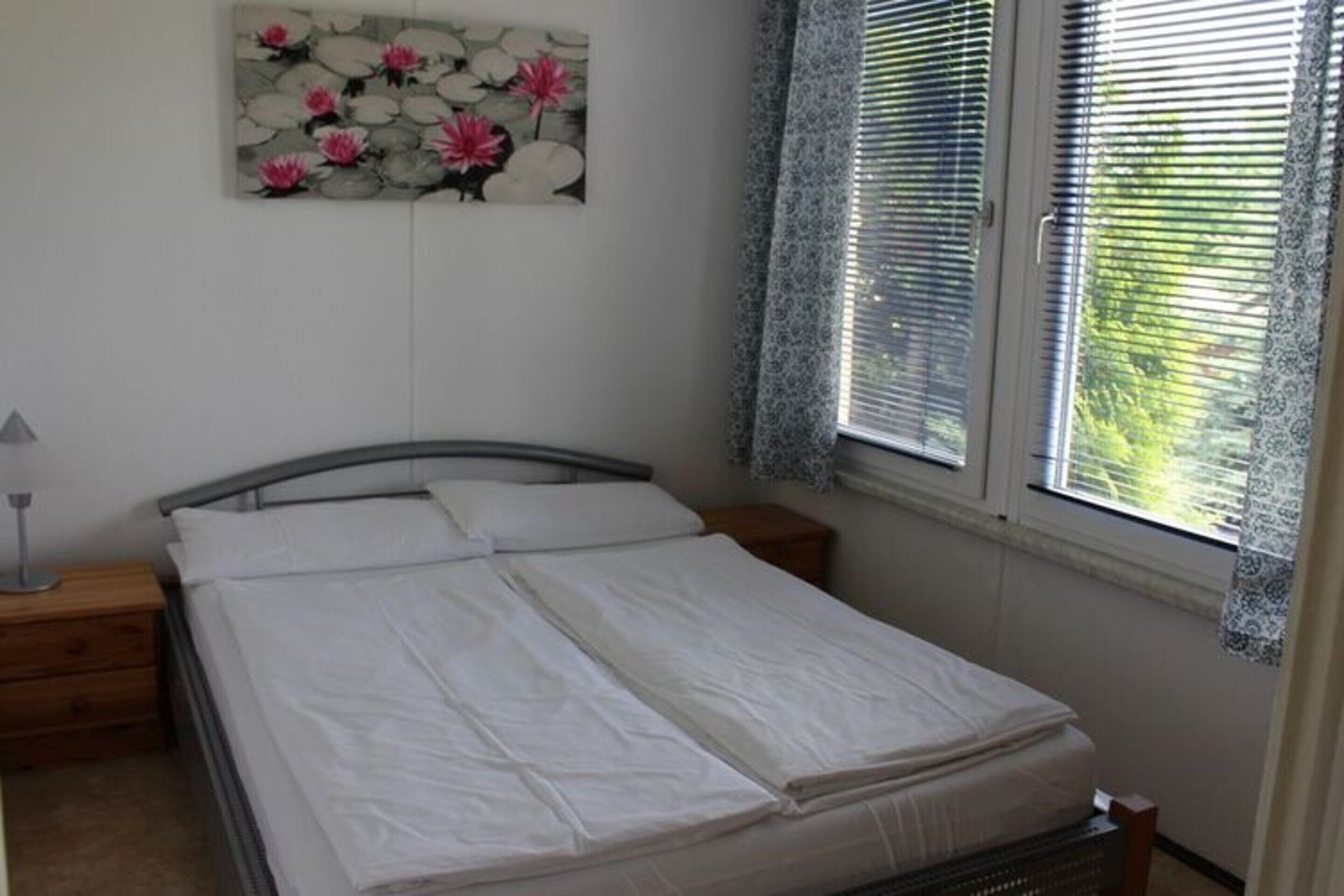 A neatly made bed in a room with a painting of flowers on the wall, a window with blinds, and a bedside table with a lamp.