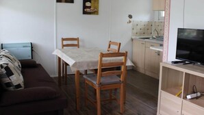 A compact living space with a dining table, two wooden chairs, a sofa, a TV on a stand, a radiator, and a kitchen area with a sink and tiled backsplash. - Bungalow Oberspreewald 3 (Schwielochsee)